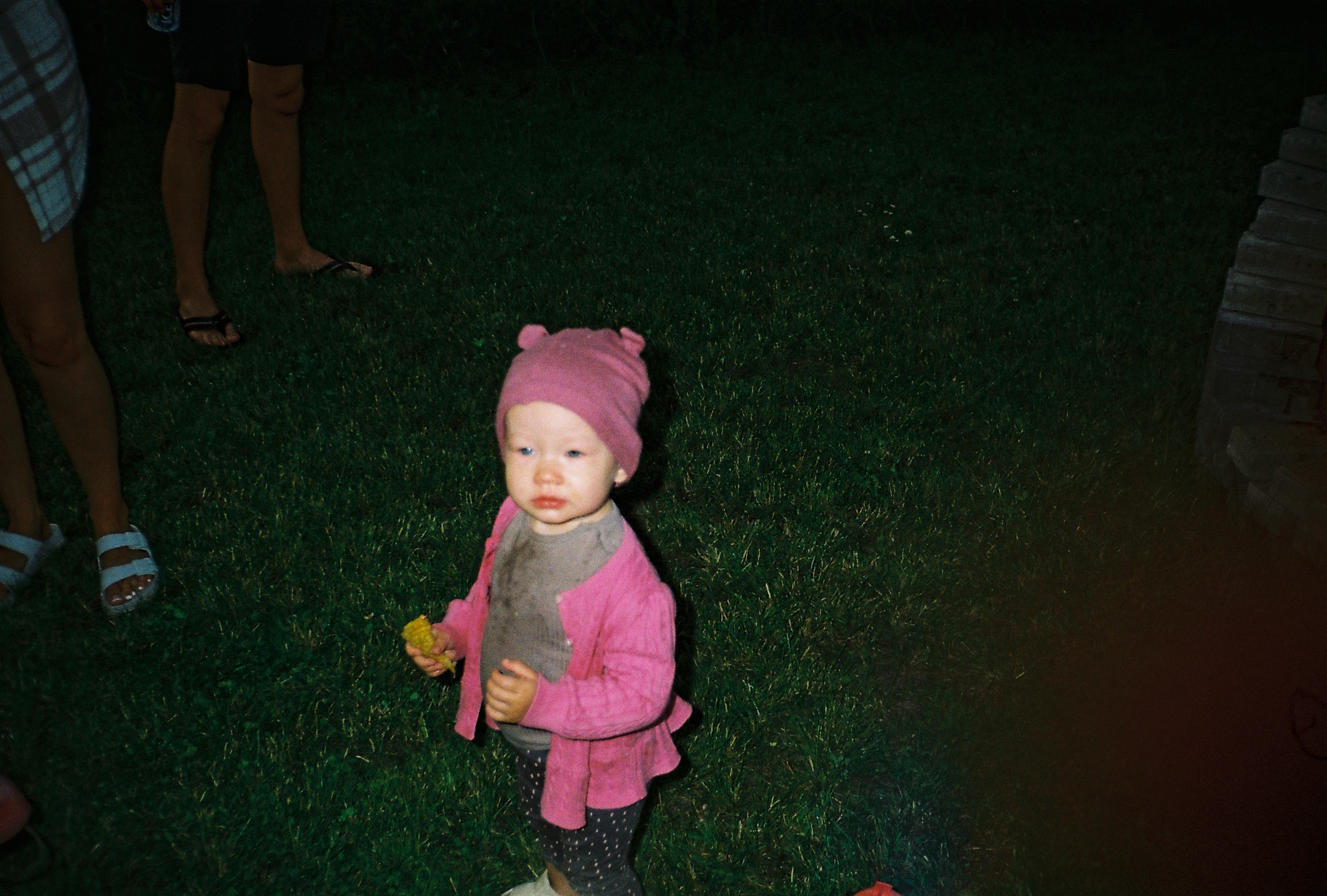 "A candid nighttime film photograph of a small child in a pink hat and sweater standing on a dark lawn, captured with a direct flash.