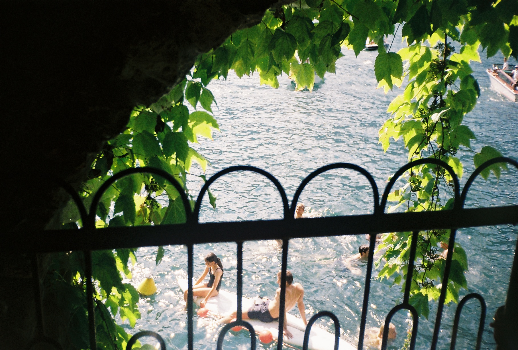 A sun-drenched film photo looking through a dark stone archway draped in green leaves toward a sparkling blue sea with people swimming.