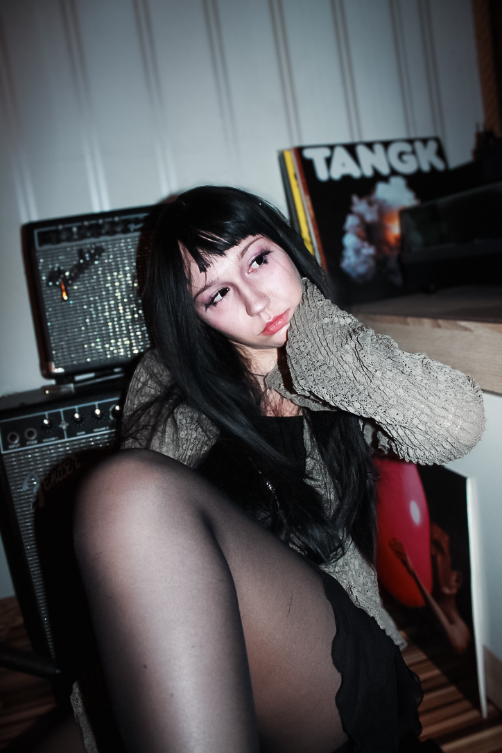 A candid-style portrait of a young woman leaning on an amplifier in a room filled with musical and artistic equipment, capturing a quiet, honest moment.