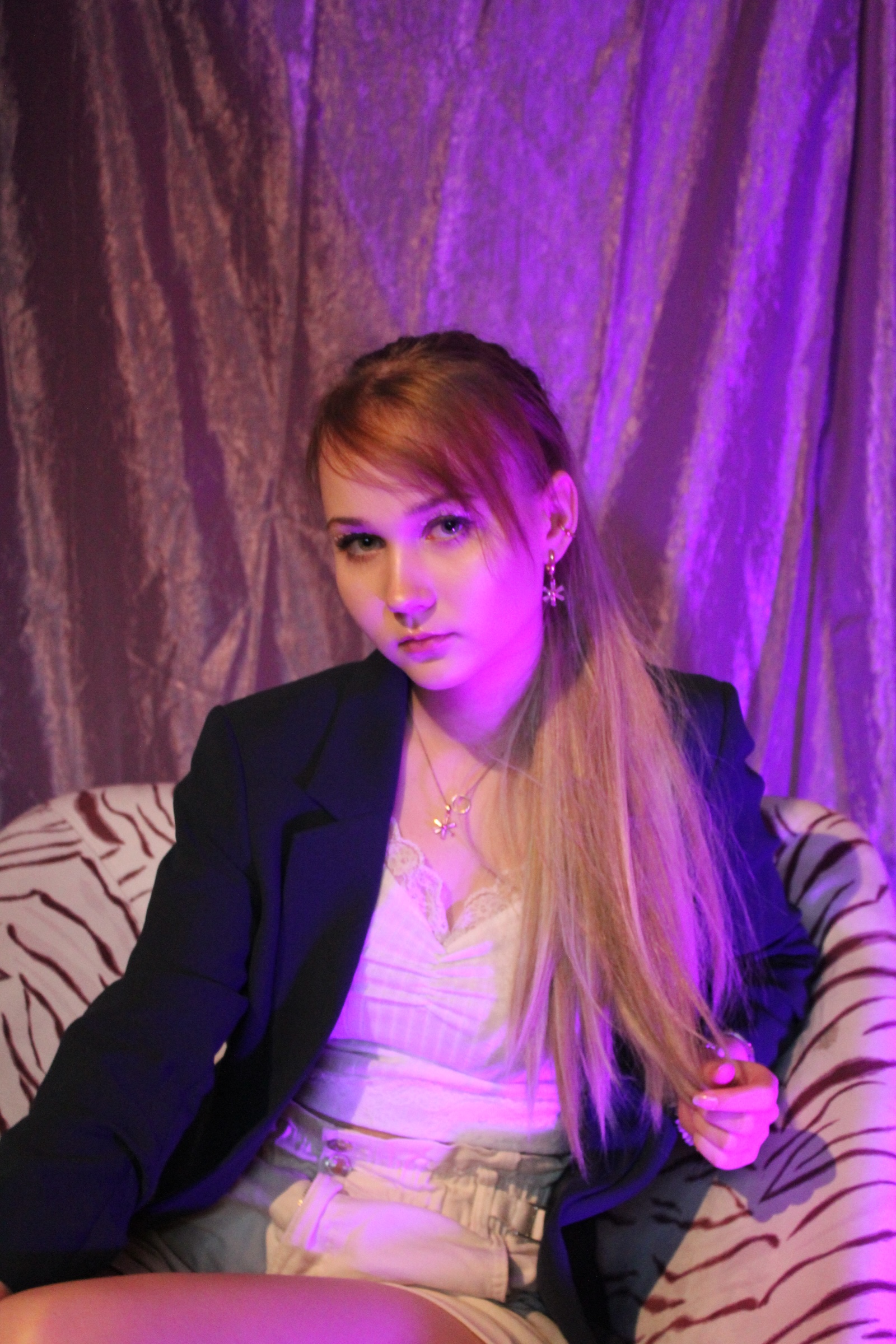 A studio portrait of a young woman with blonde hair sitting in a zebra-patterned chair, illuminated by vibrant blue and purple neon lighting against a draped curtain background.