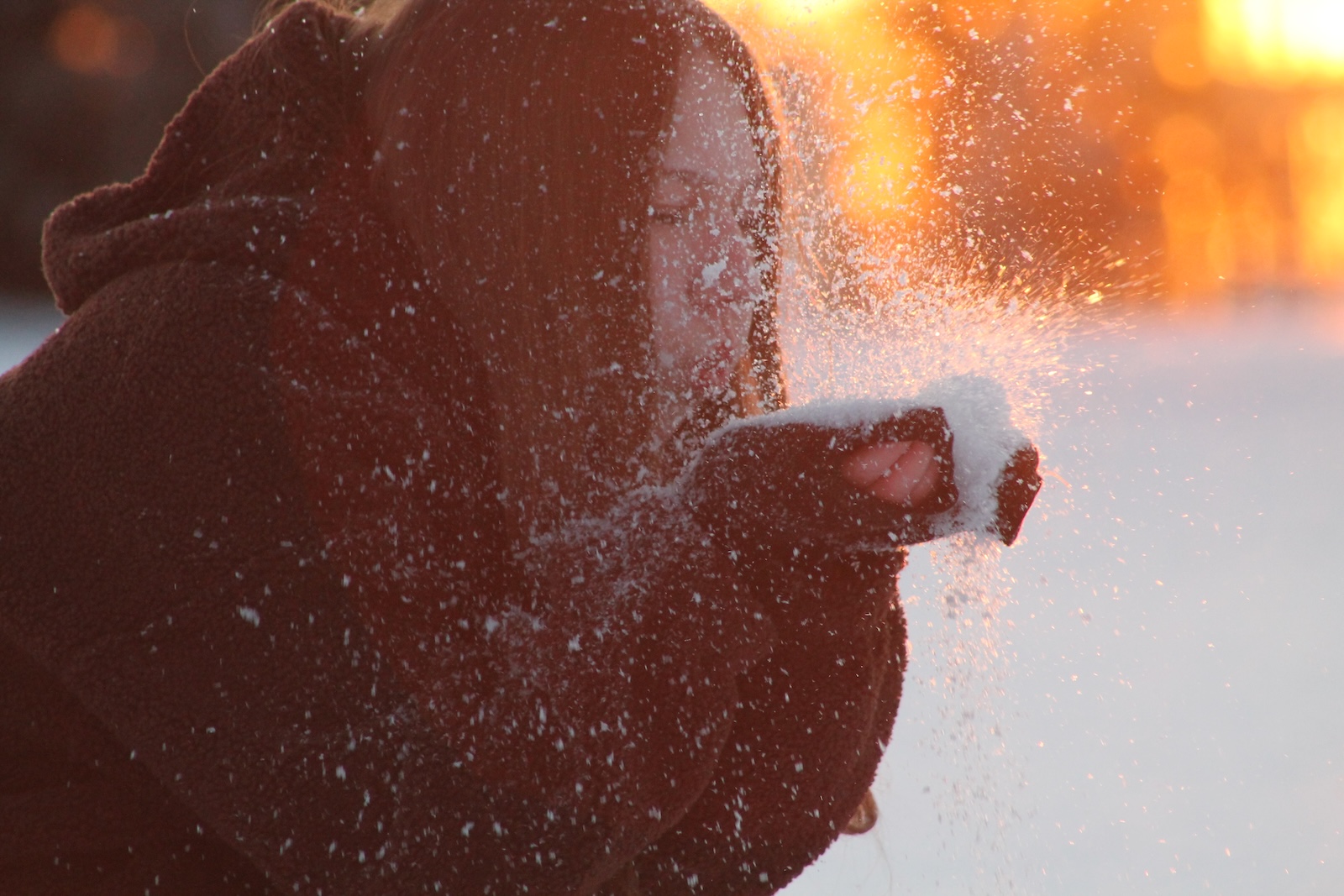 A close-up artistic shot of a person blowing snow from their hands, with bright golden hour sunlight creating a sparkling effect.