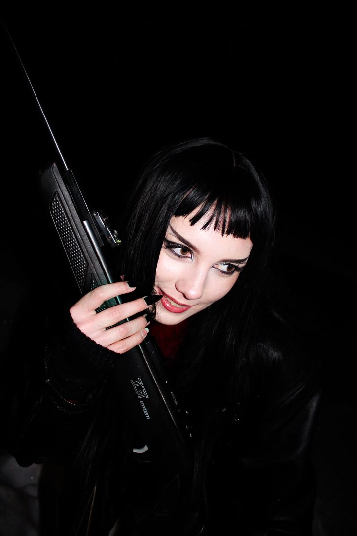 A moody, high-contrast portrait of a young woman with dark hair and bangs holding a prop against a black background, emphasizing composition and shadow.
